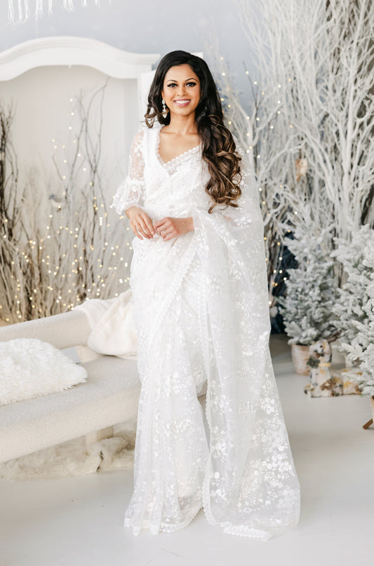 Model draped in a white transparent mesh saree with white floral embroidery and a white lace border. Model is wearing a matching white blouse with flared sleeves and a white saree petticoat underneath. Christmas themed background.