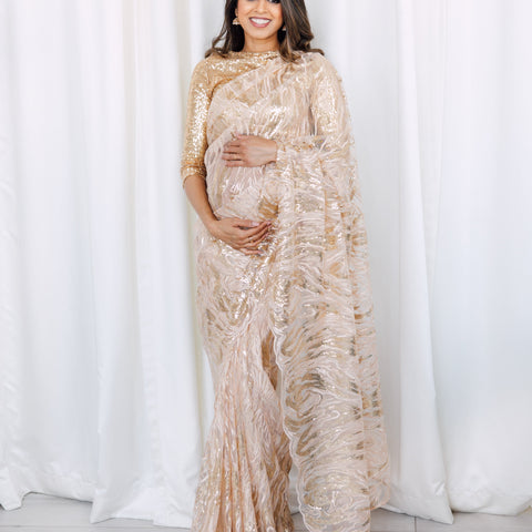 Model holding her baby bump while draped in a gold mesh saree embroidered with sequins and finished with a gold thread scalloped edge. Model is also wearing a high neck light gold sequin crop top with 3/4 length sleeves.