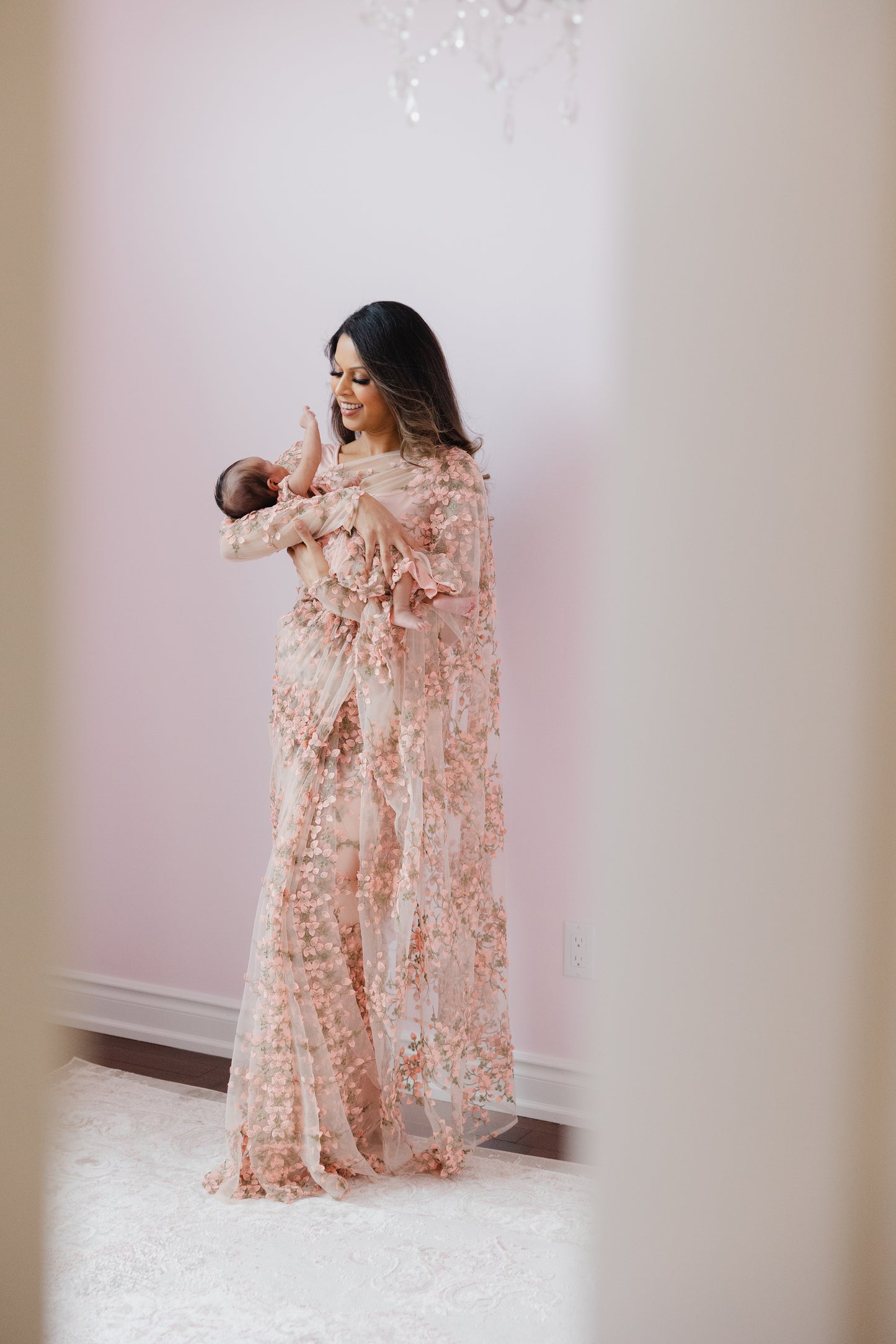 Model holding a baby. Model is draped in a blush pink transparent saree with bright pink floral embroidery and matching long sleeve blouse.