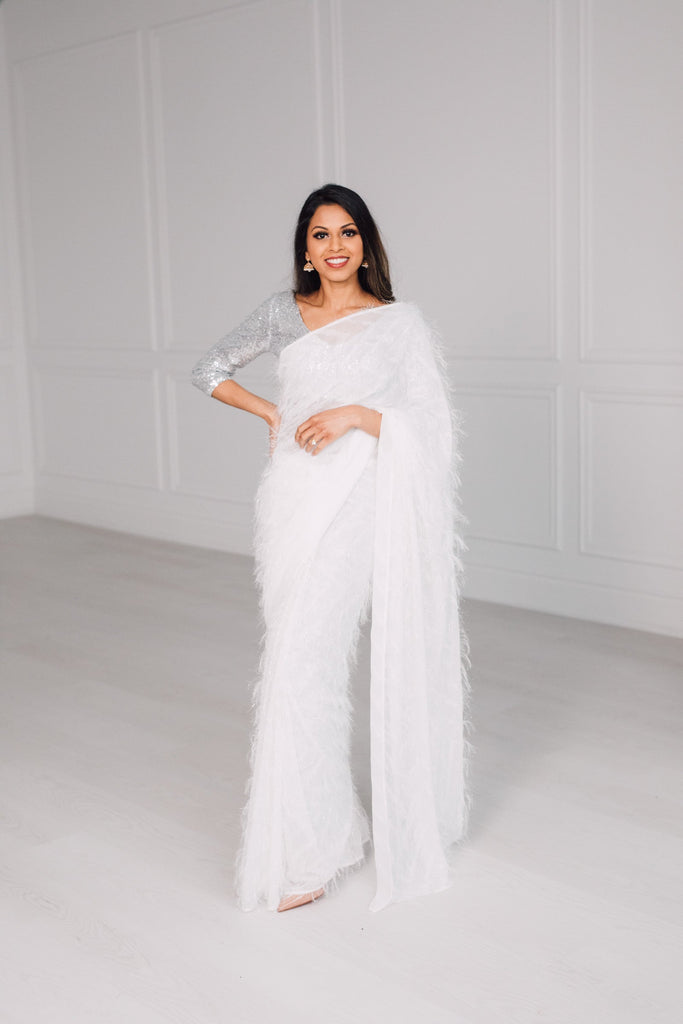Model draped in a white saree featuring threadwork that creates the illusion of 3D feathers. Model is also wearing a silver v-neck 3/4 sleeve sequin crop top.