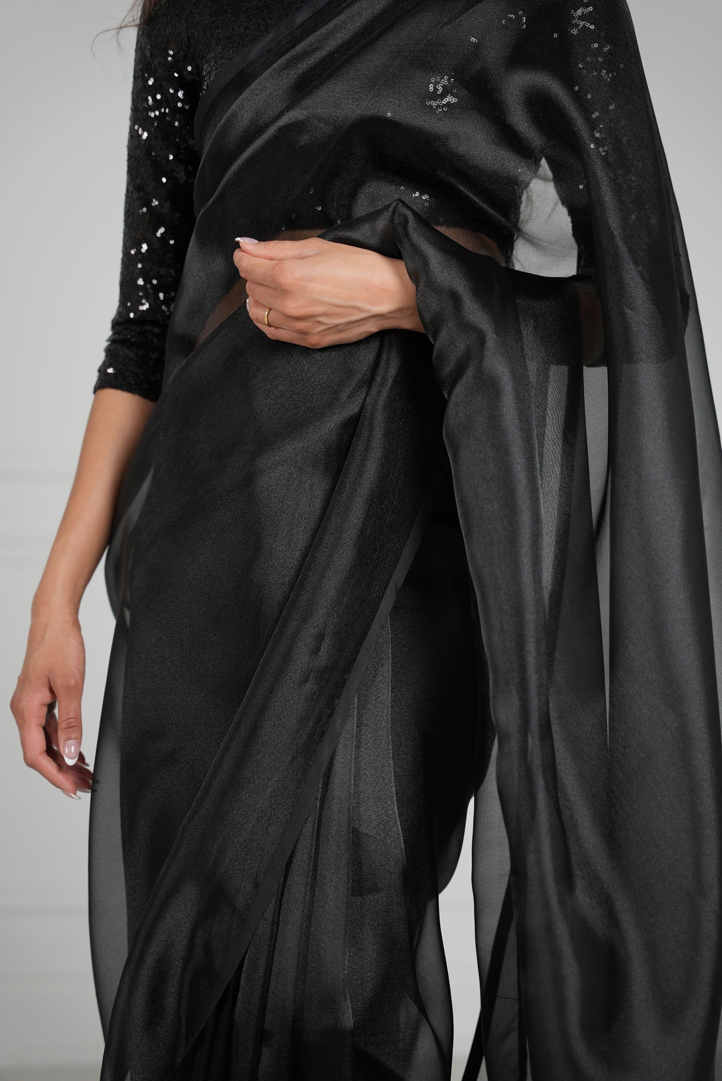 Model draped in a black organza saree with black satin border. Model is also wearing a black 3/4 sleeve sequin crop top.