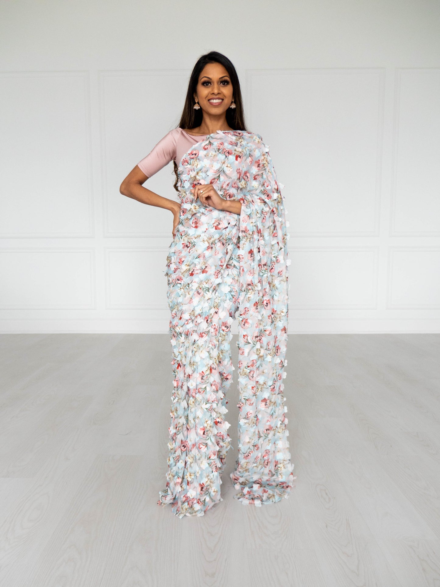 Model draped in a lightweight pale blue saree with a red, green, and pink floral print and 3D petals. Model is also wearing a blush pink half sleeve crop top.