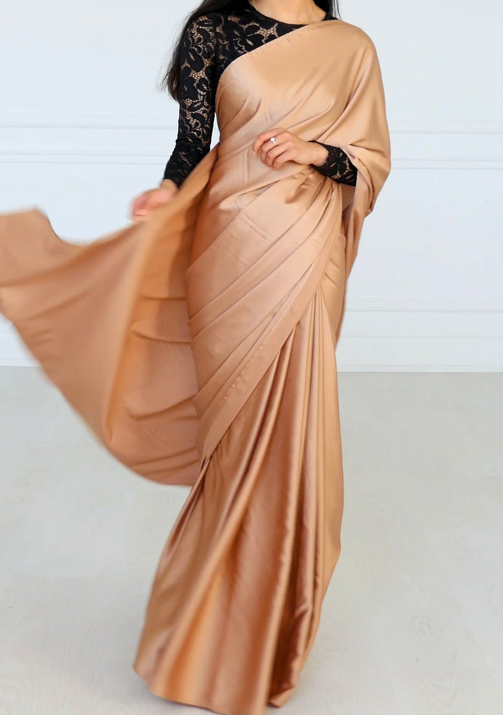 Model wearing a black long sleeve lace crop top and draped in a champagne colored satin silk saree. Model is swinging her pallu.